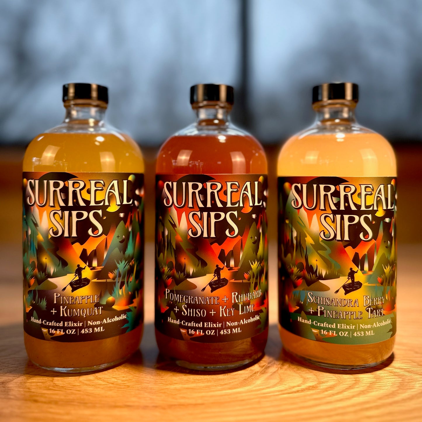 Three Surreal Sips hand-crafted non-alcoholic elixir bottles lined up on a wooden table