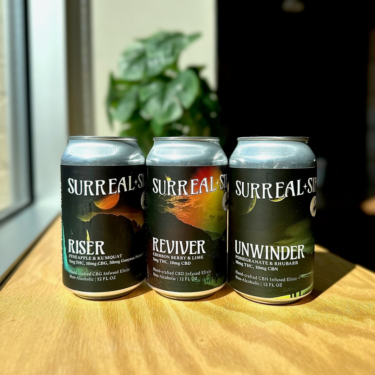 Three Surreal Sips THC seltzer cans — Riser, Reviver, and Unwinder — on a wooden table